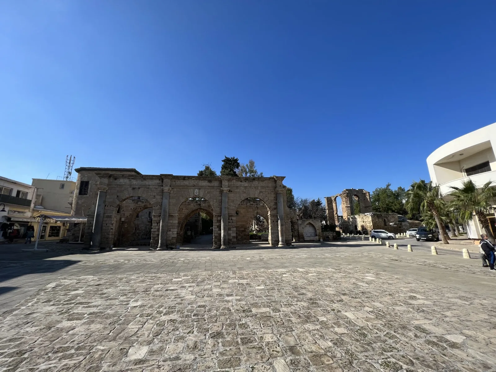 Wide-angle view of a historic stone archway with multiple arches under a clear blue sky, surrounded by modern buildings and a cobblestone plaza.