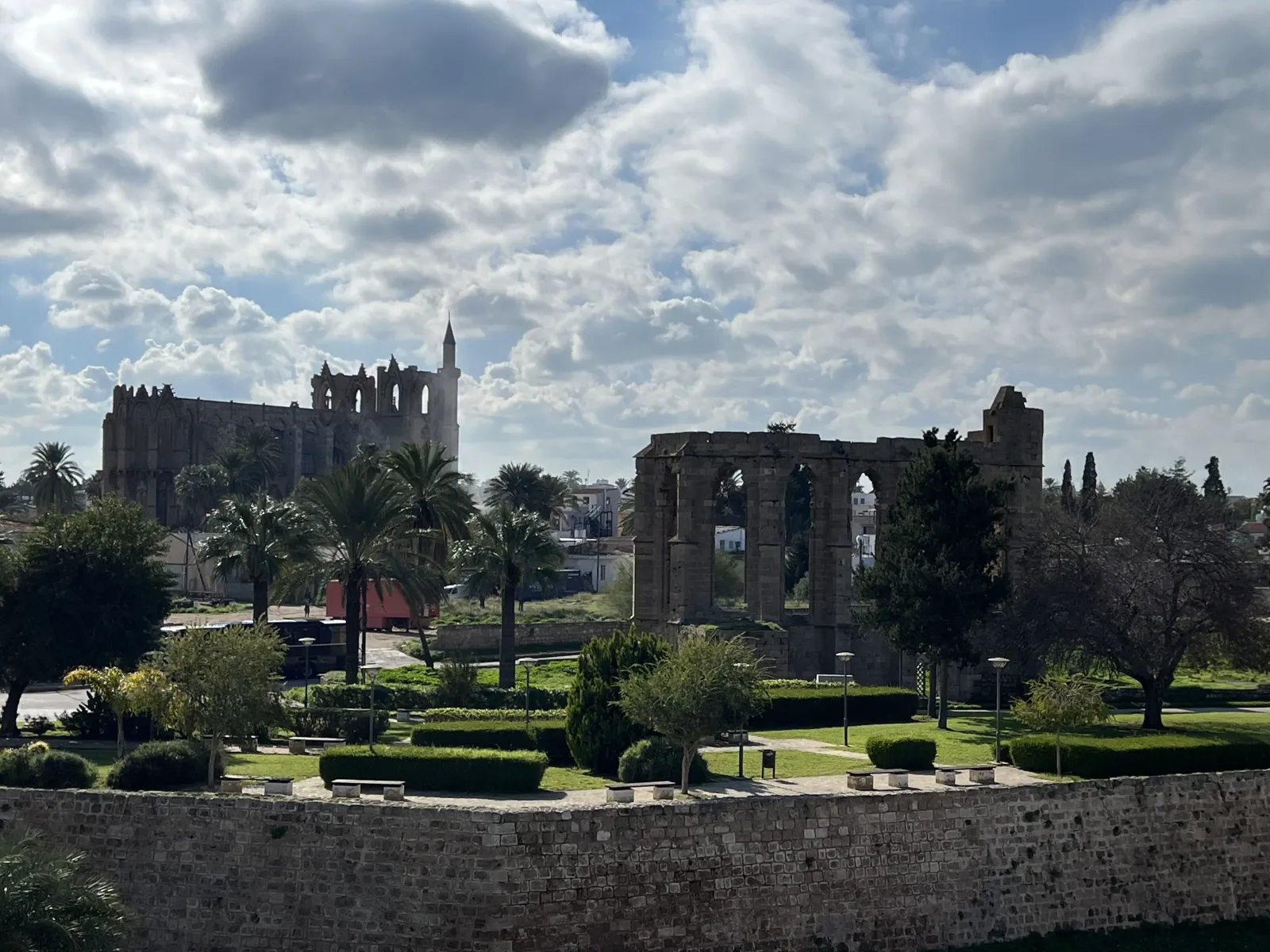 Partly cloudy sky over ancient stone ruins with arched doorways, surrounded by manicured lawns and modern buildings in the background.