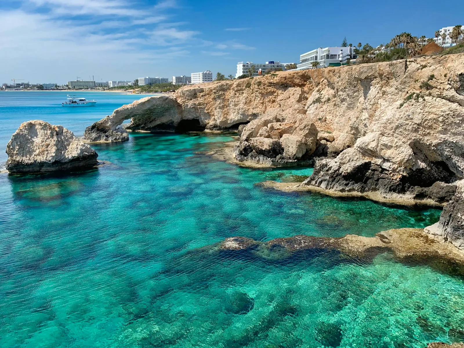 A stunning coastal view featuring crystal clear turquoise waters, rocky formations, and a view of a beachfront city in the distance.